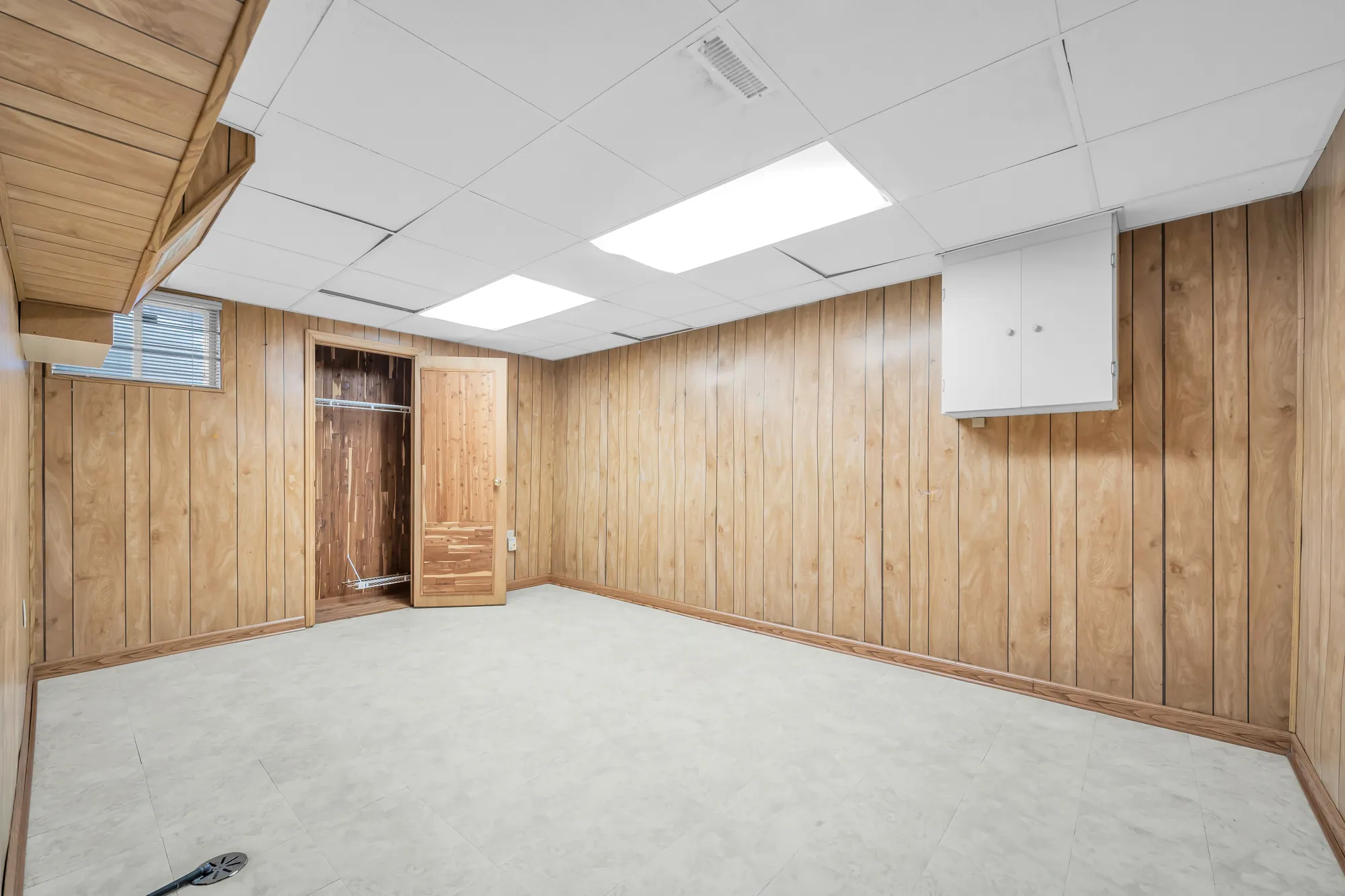 A basement room with wood-panelled walls, a dropped ceiling with light panels, a closet with its doors open, a small window