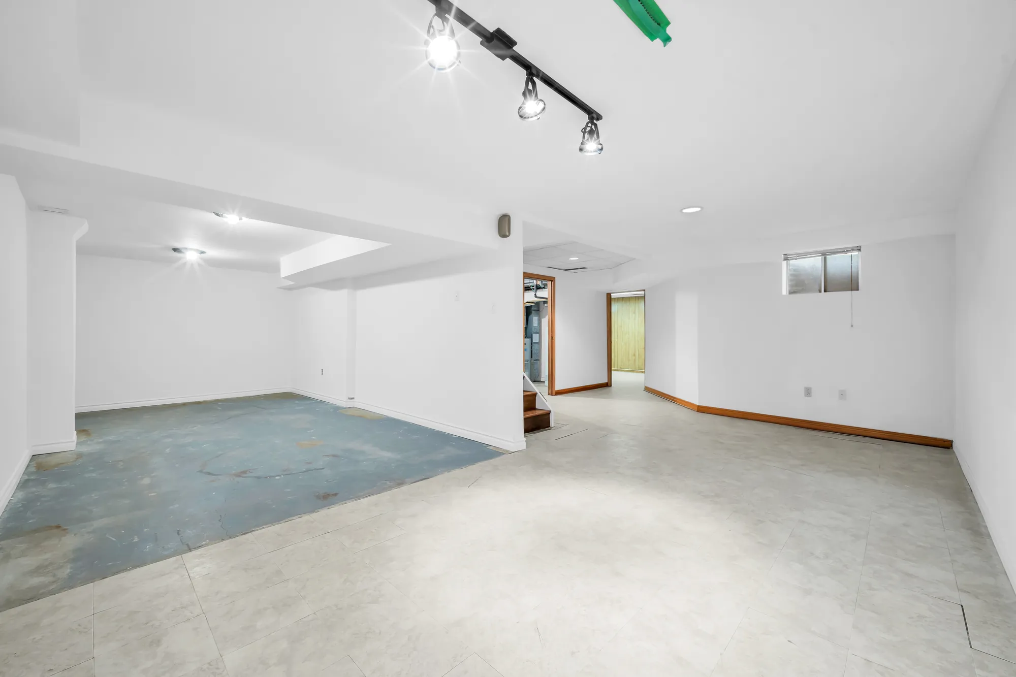 The open part of the basement: white walls, pot lights and track lights, a painted floor, small windows, the stair beyond