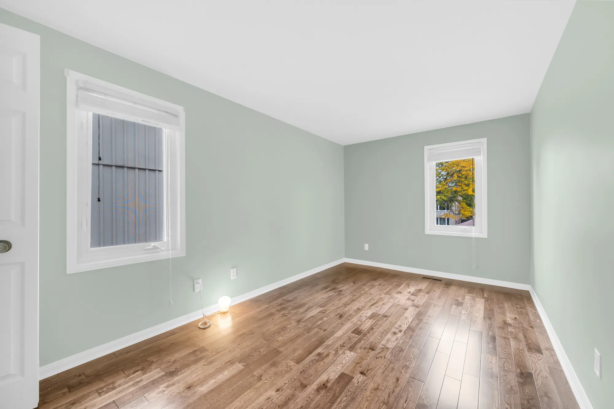 A second bedroom with sage green walls, empty: a window on the side wall, another at the end, wood floors
