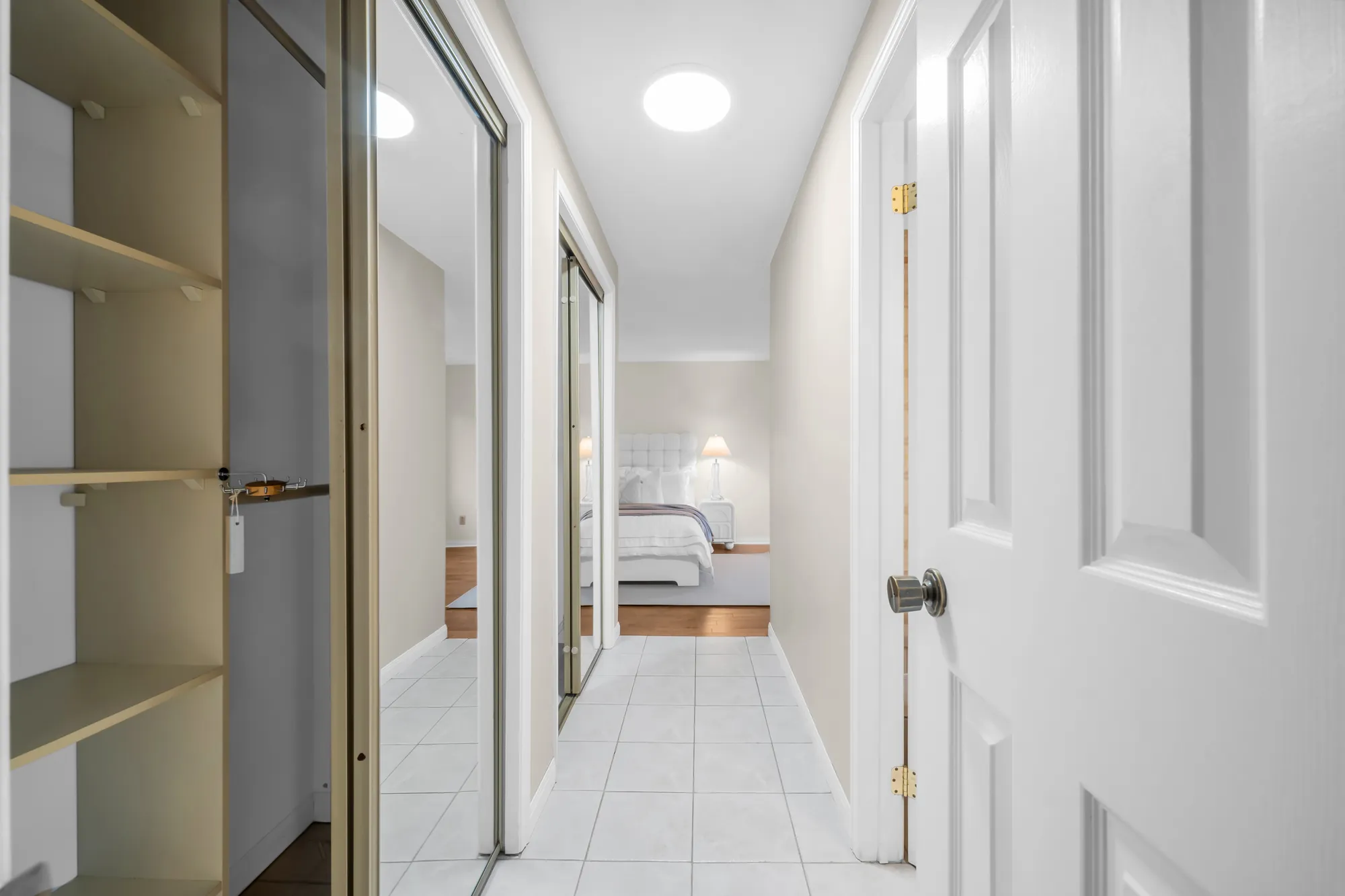 A passage lined with closets: mirrored sliding doors on one side, open shelving on the other, a tiled floor, the bedroom beyond