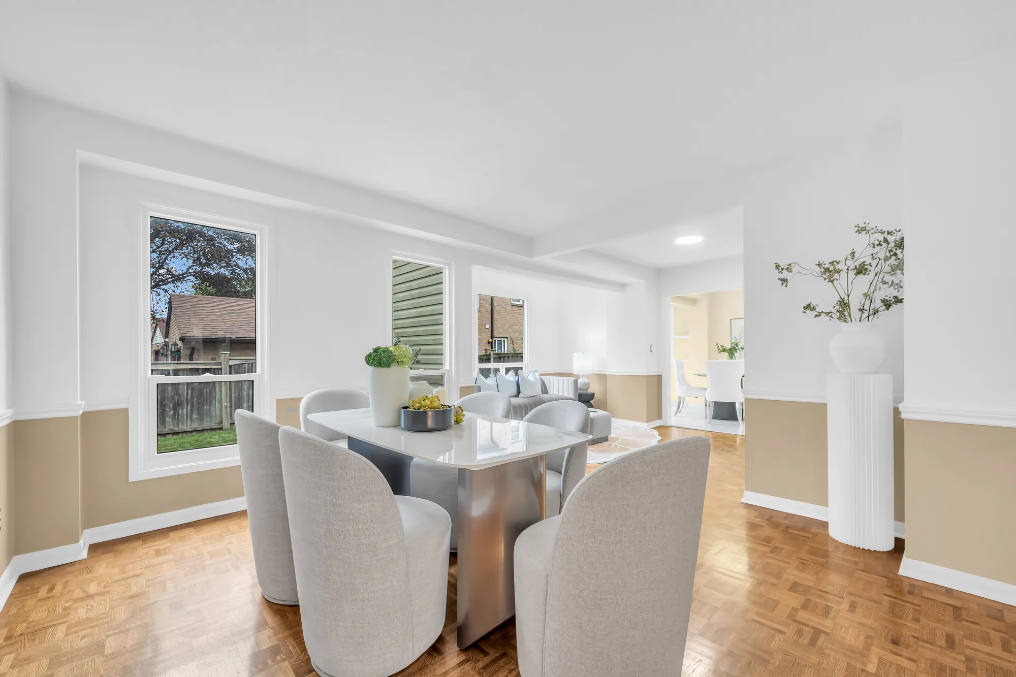 The dining table toward the windows over the yard, the sitting area beyond, a white pedestal with branches