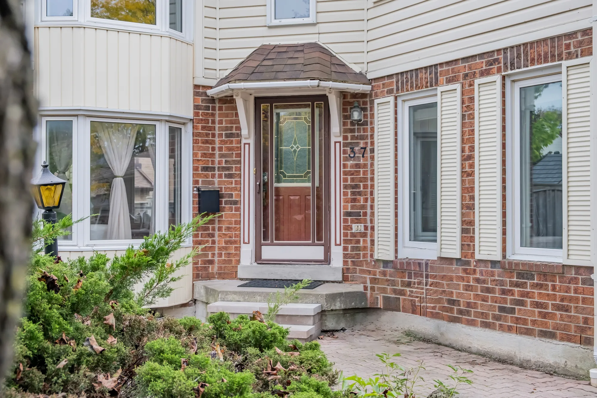 The front door under a small shingled hood: a door with a leaded-glass panel behind a storm door, the number 37 on the brick beside it, the bay window at the left, shrubs in front