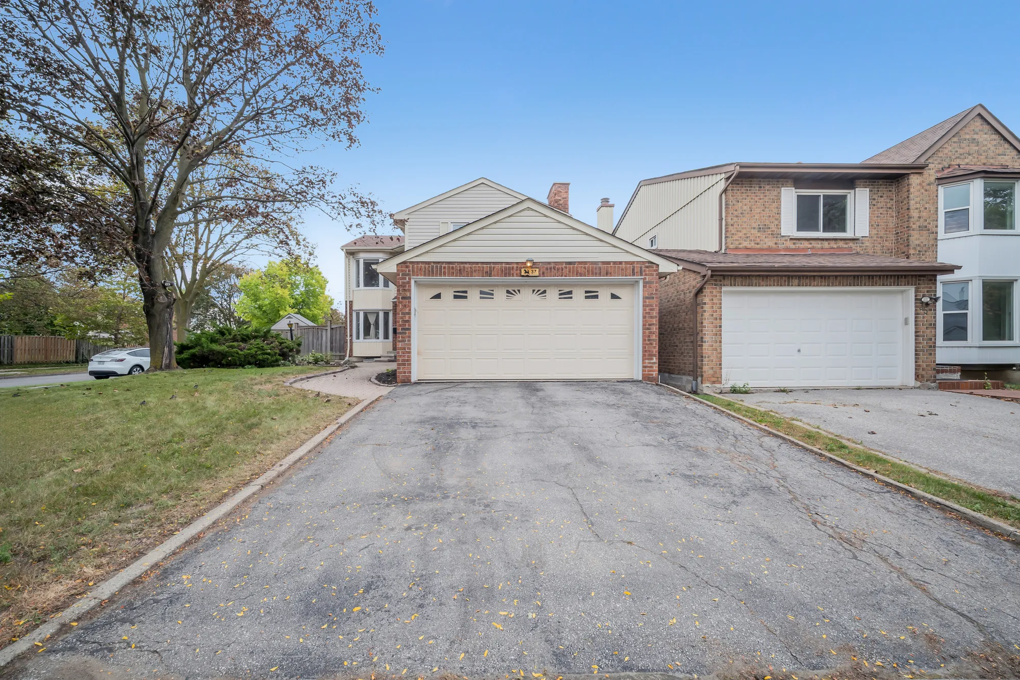 The drive and the double garage from the street, a house number plaque over the garage door, the neighbouring house at the right, a tree and lawn at the left