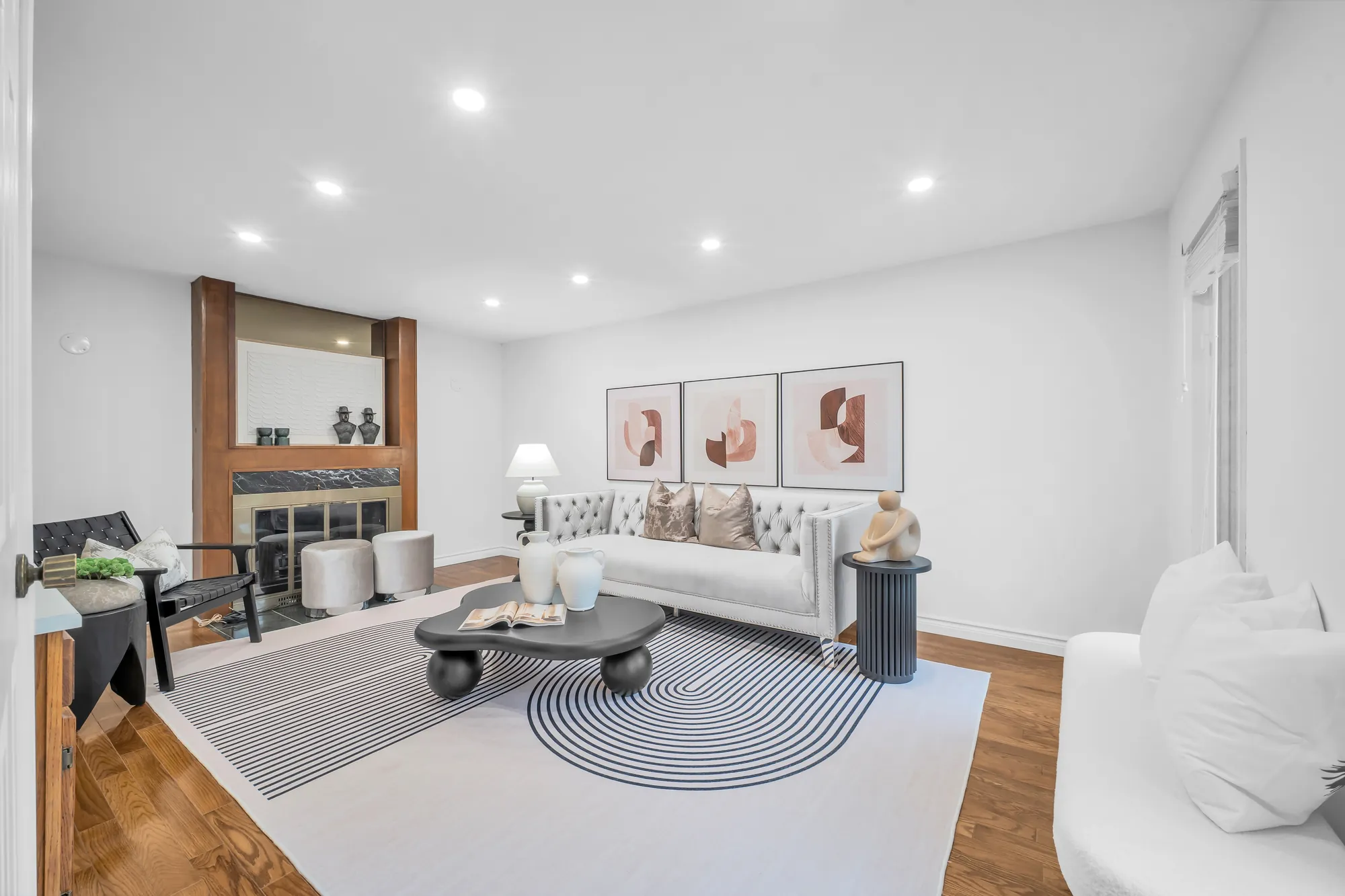 The family room: a fireplace in a wood surround, pot lights, wood floors, a tufted sofa, a lounge chair and a round coffee table on a striped rug