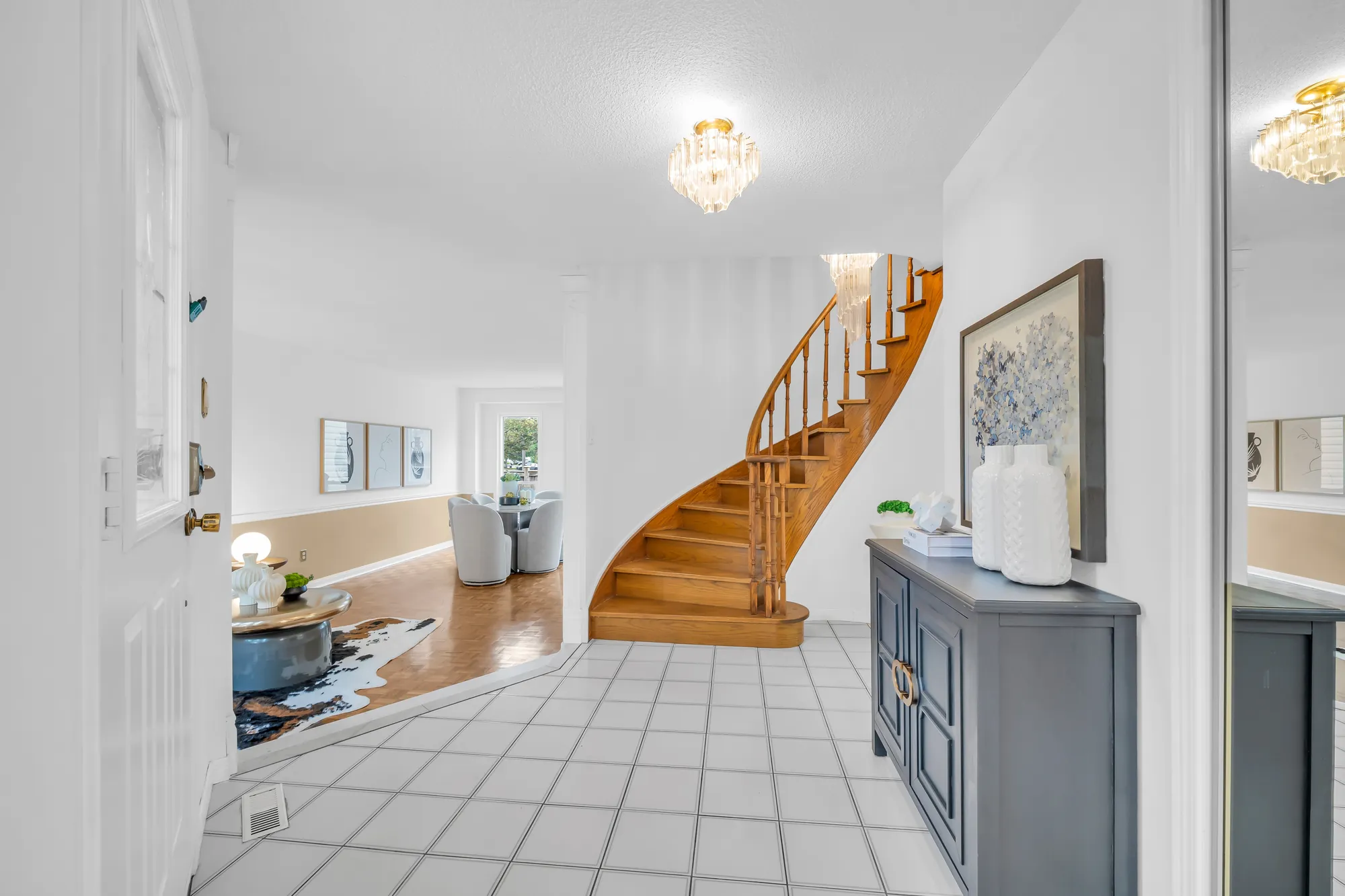 The foyer toward the curved oak staircase, a crystal light overhead, a grey cabinet at the right, the living room at the left