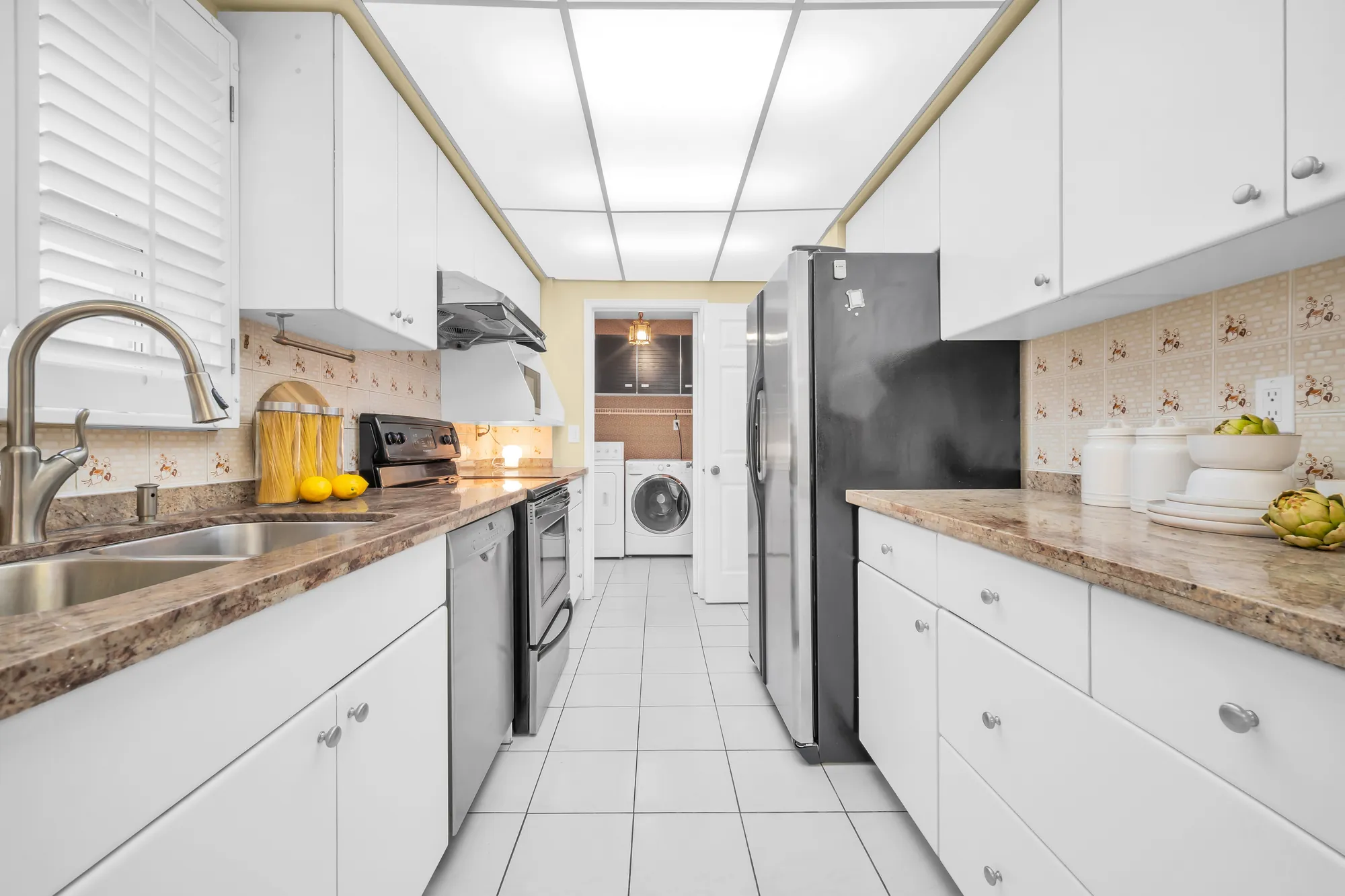 The kitchen: white cabinets on both sides, stone-look counters, a double sink under a shuttered window, a stainless fridge, range and dishwasher, the laundry room through the doorway at the end