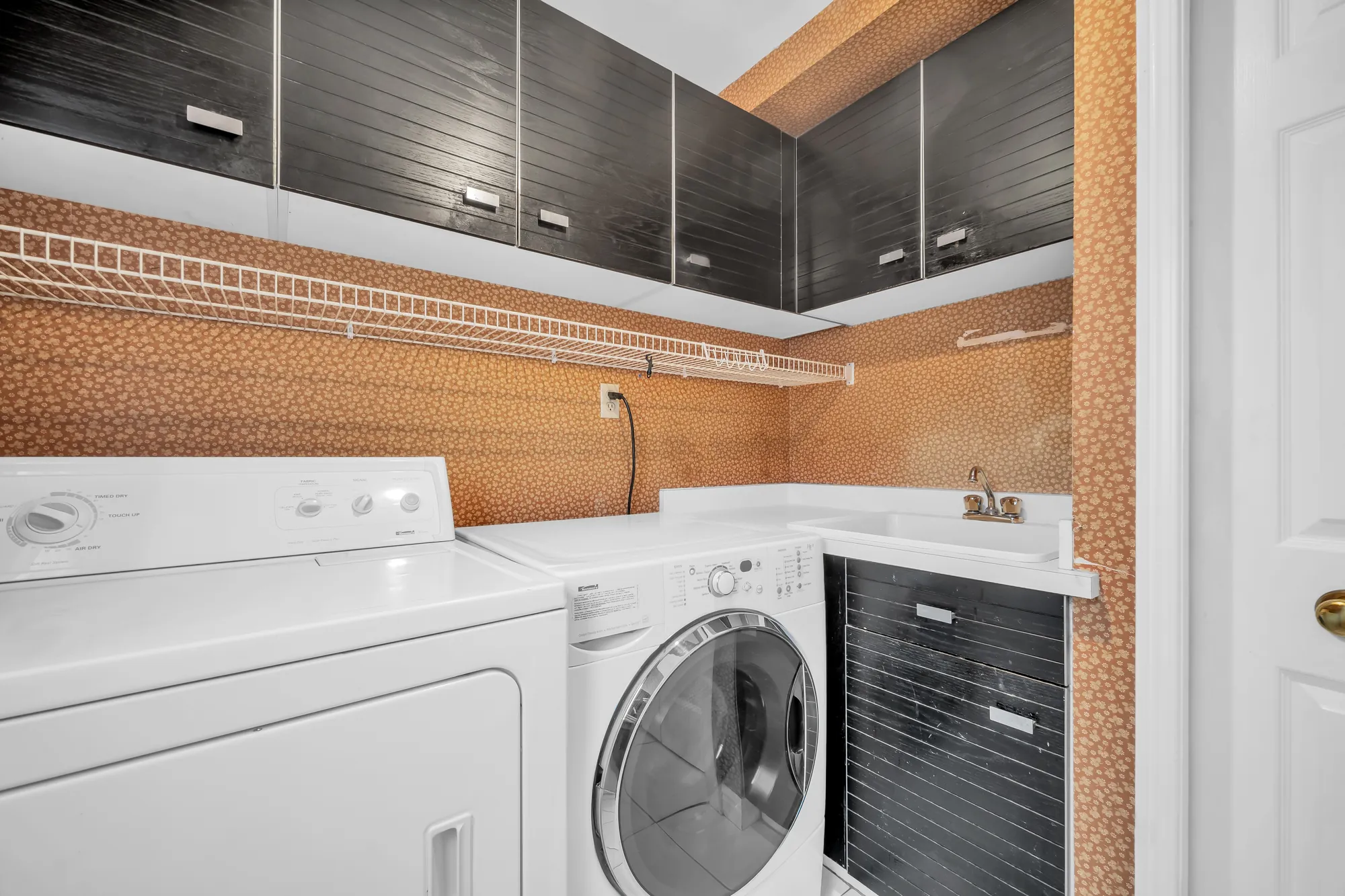 The laundry room: a washer and a dryer, a laundry sink, dark upper cabinets, a wire shelf, patterned wallpaper