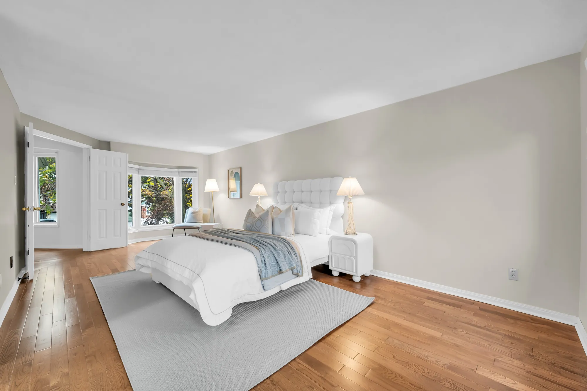 The primary bedroom: a bed with a white tufted headboard, two night tables and lamps, a grey rug, wood floors, a bay window at the far end