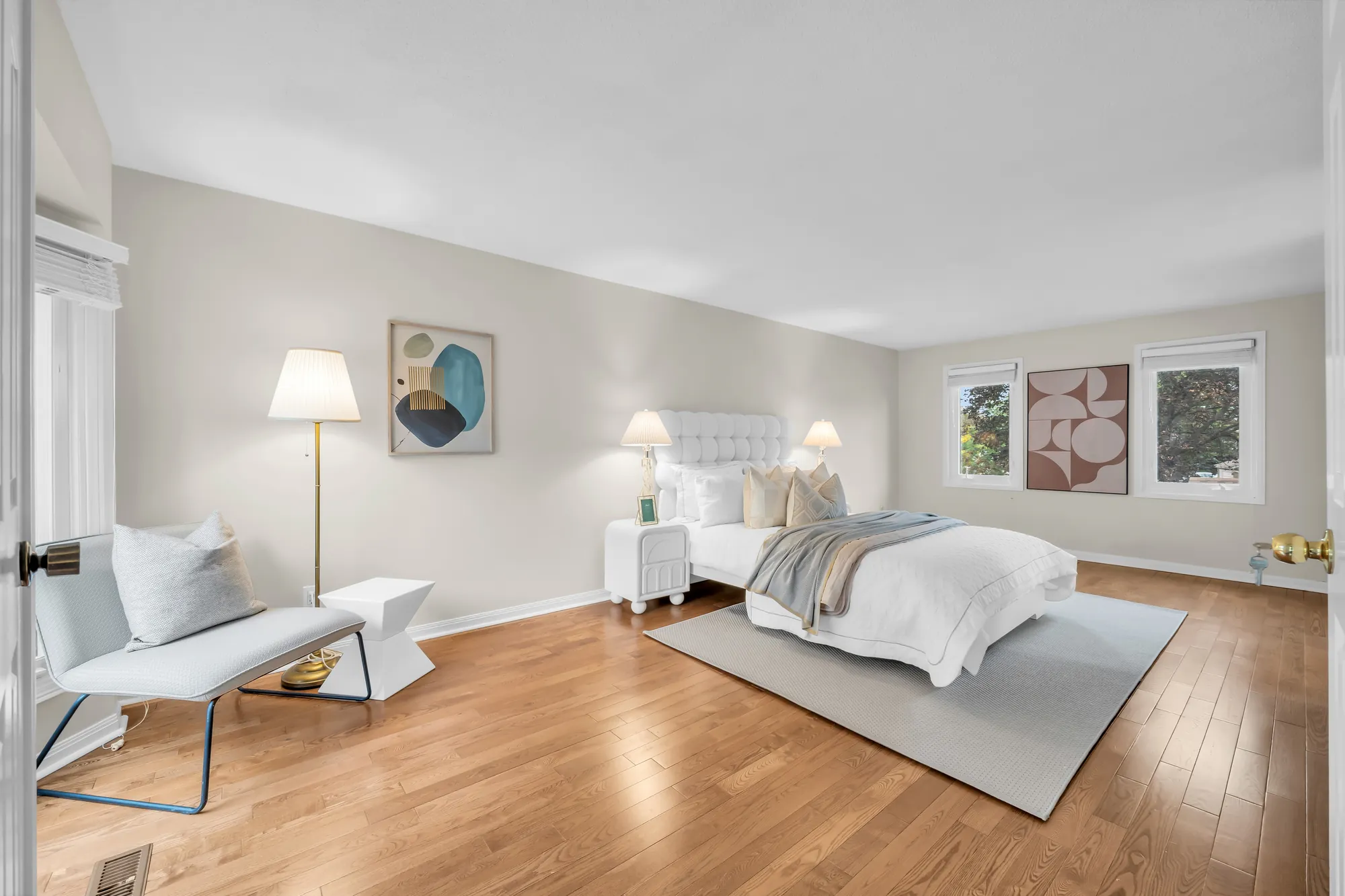 The primary bedroom toward its windows: the bed, a floor lamp and a lounge chair, framed prints