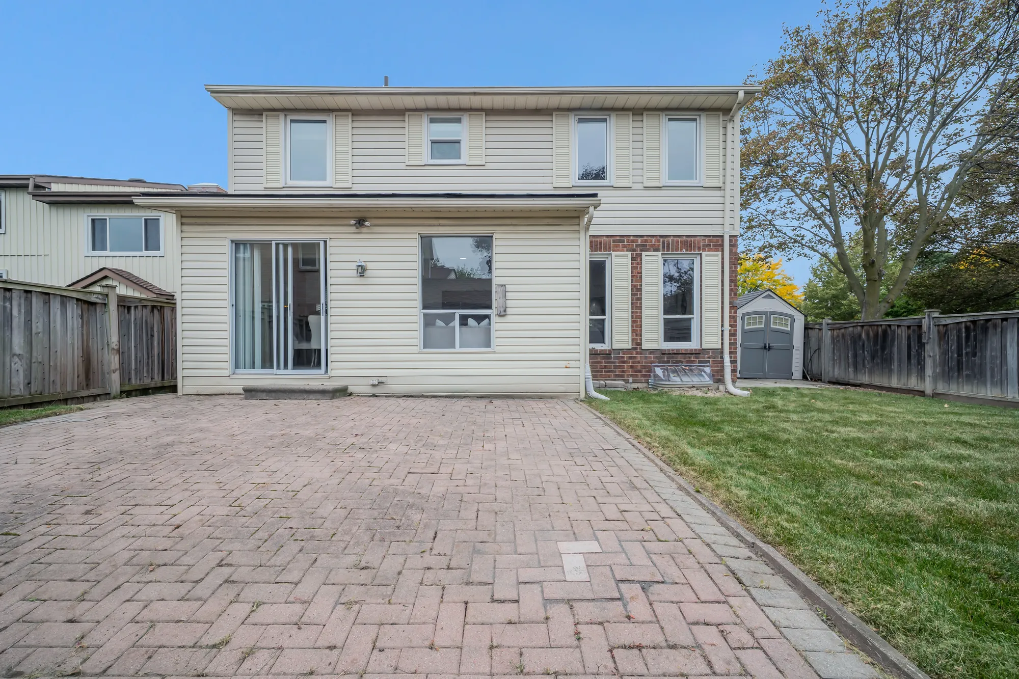 The back of the house by day: a single-storey addition with a sliding glass door and a window, two storeys above it, an interlocking brick patio, a lawn, a garden shed and a fence