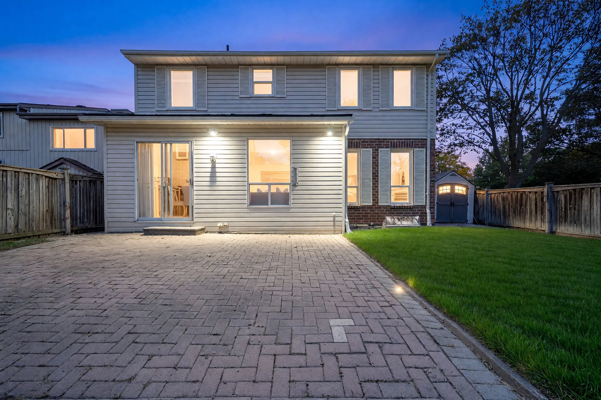 The back of 37 Wintermute Boulevard at dusk: two storeys in cream siding and red brick, a single-storey addition with a sliding glass door, the windows lit, an interlocking brick patio and a lawn